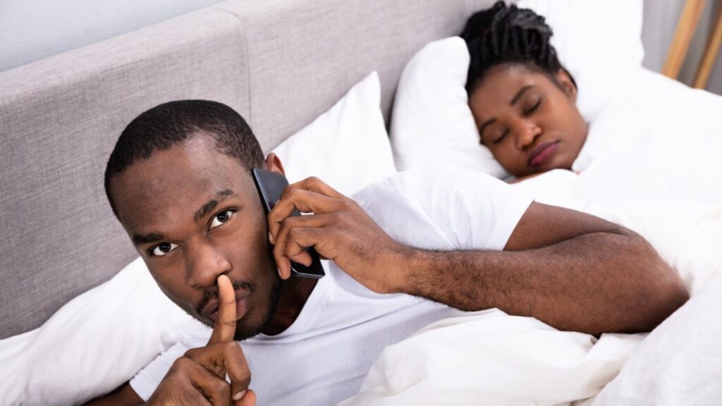 man in bed showing shh on the phone while woman sleeps next to him