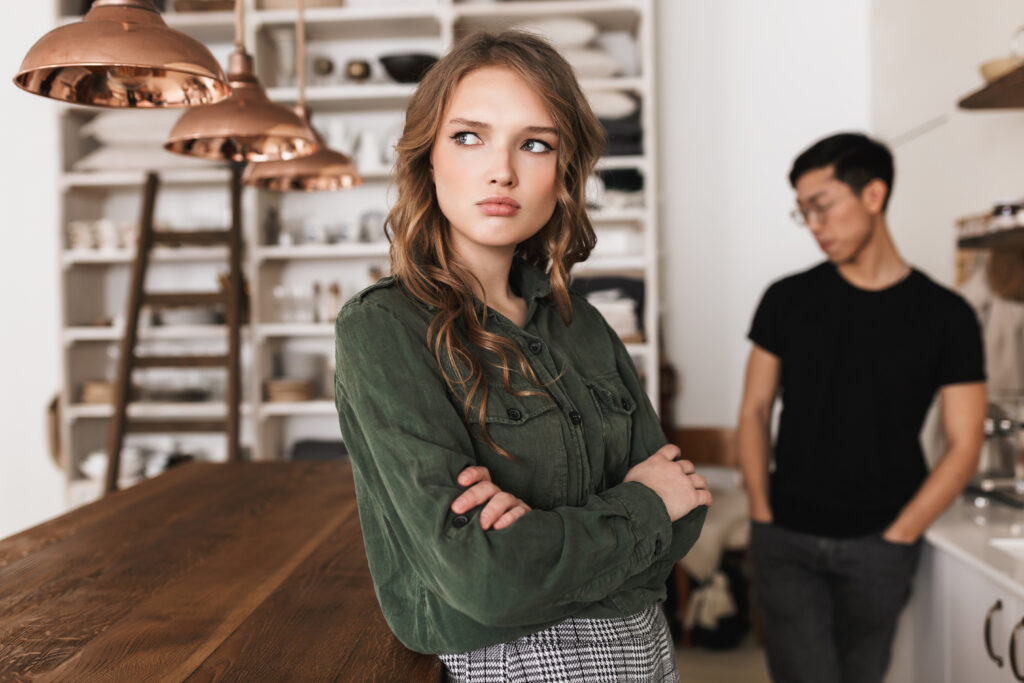 Attractive upset woman with wavy hair holding hands together sadly looking aside with thoughtful asian man on background. Young international couple quarreling spending time on kitchen at home